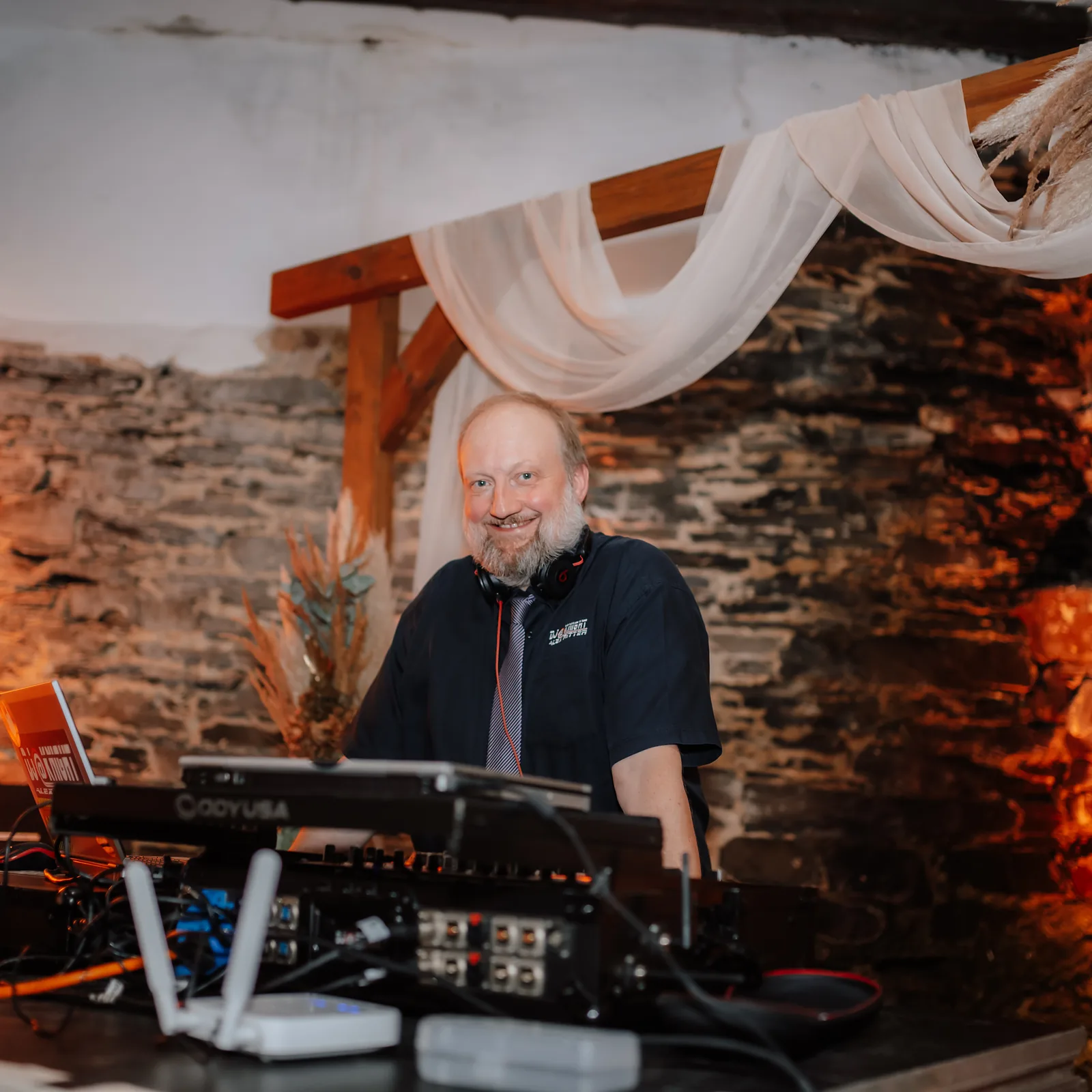 DJ Alex Ritter von DJ@Knight lacht hinter seinem DJ-Pult, &uuml;ber ihm h&auml;ngen helle Stoffbahnen und Trockenblumen des Traubogens.