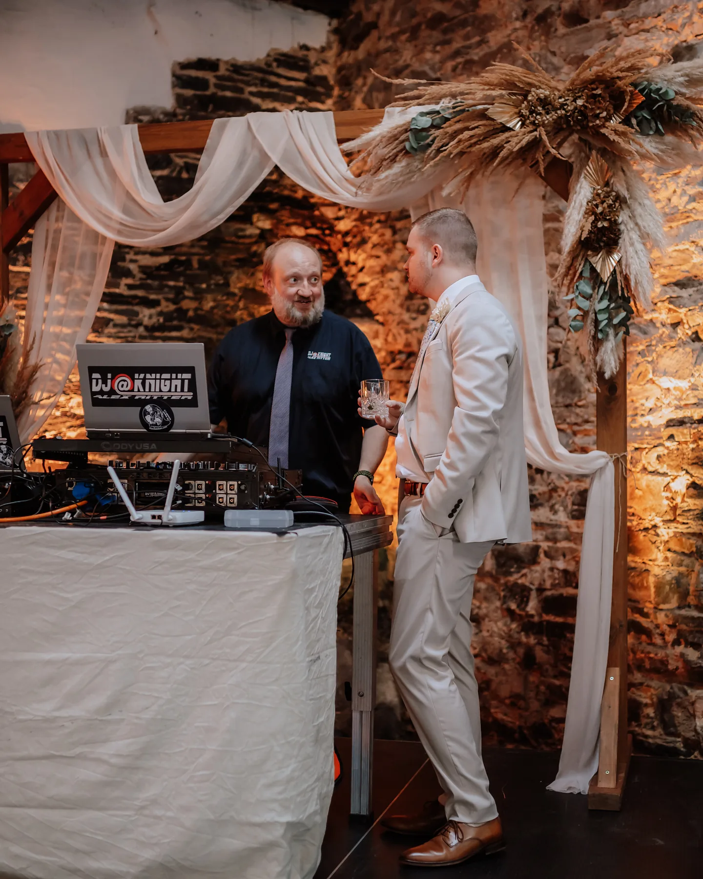 DJ Alex Ritter von DJ@Knight im Gespr&auml;ch mit einem Hochzeitsgast am DJ-Pult unter dem geschm&uuml;ckten Traubogen.