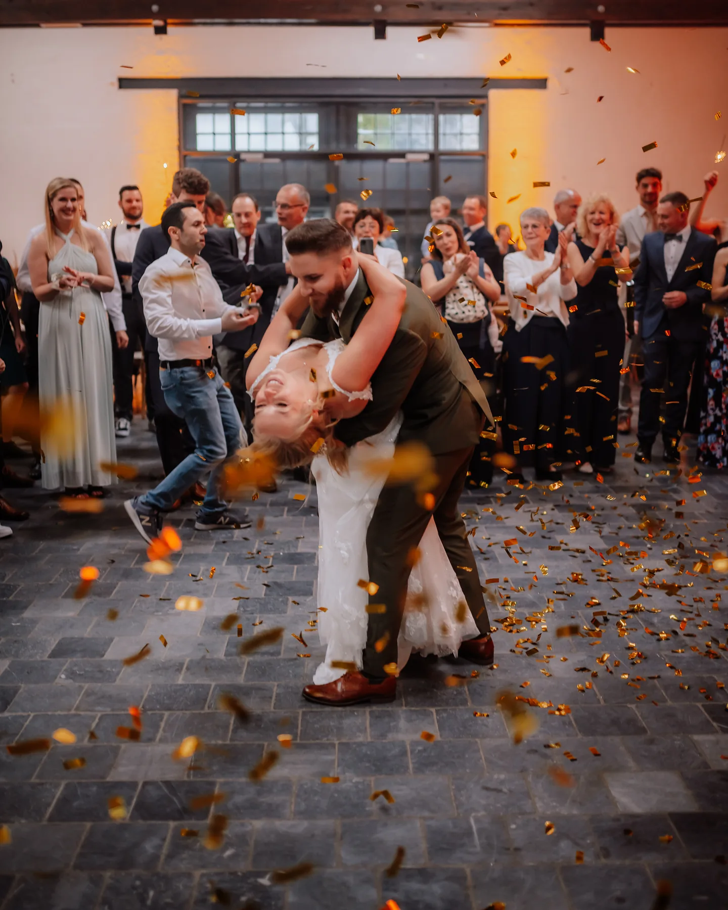 Der Br&auml;utigam l&auml;sst die Braut beim Hochzeitstanz im goldenen Konfettiregen nach hinten fallen, die G&auml;ste stehen klatschend im Kreis.