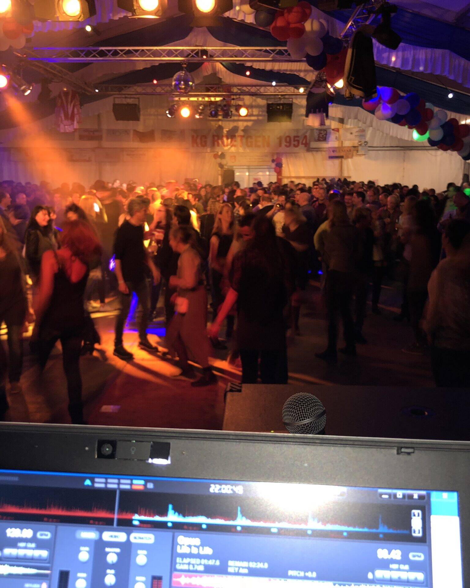Volle Tanzfl&auml;che im Festzelt in orangem Licht, vorn der Notebook-Bildschirm am DJ-Pult, hinten das Banner der KG Roetgen